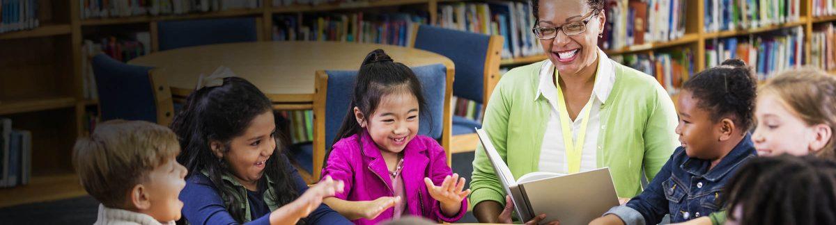 A multi-ethnic group of seven children, 6 and 7 years old, sitting at a round table in the library with their teacher, a mature African-American woman, who is reading a book to them. The boys and girls are playfully slapping the table with their hands.